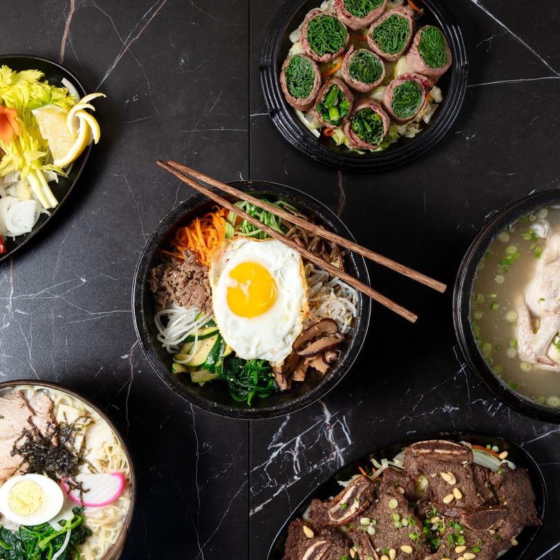 Overhead view of five Korean dishes, including bibimbap, ramen, and short ribs, on a black marble table.