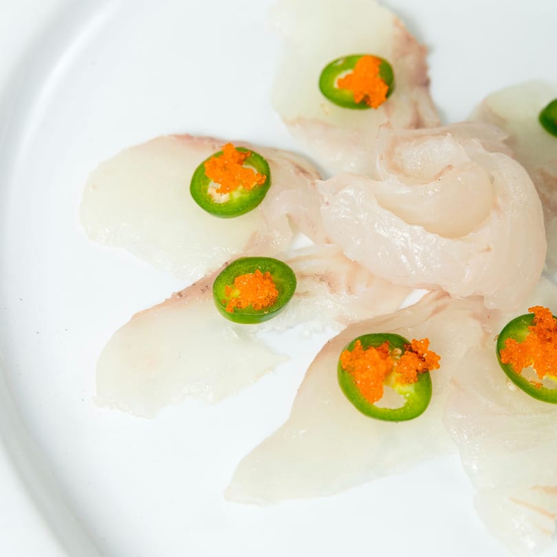 Thinly sliced white fish sashimi topped with green jalapeño slices and orange tobiko on a white plate.