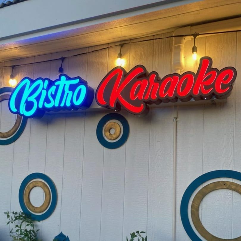 Neon signs for "Bistro" in blue and "Karaoke" in red on a white wooden wall.