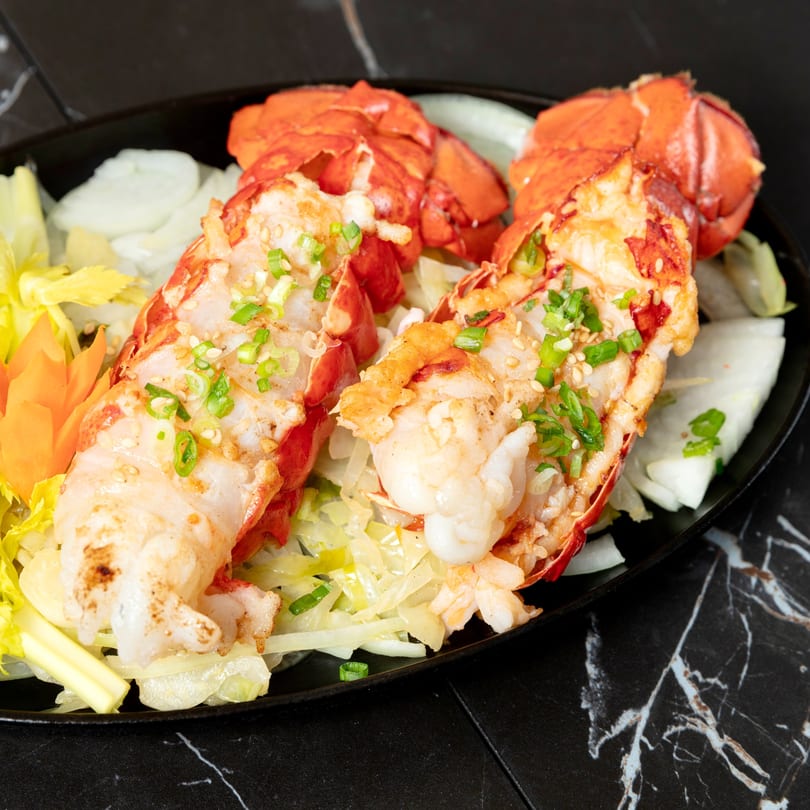 Two grilled lobster tails with green onions and sesame seeds, served on a bed of vegetables.
