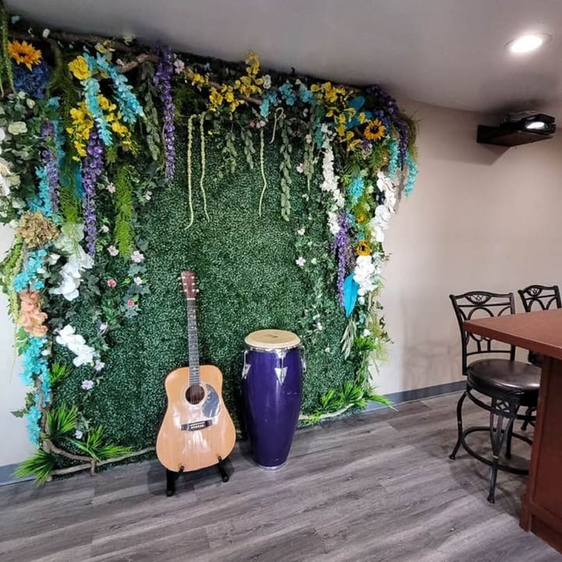 Acoustic guitar and purple drum in front of a green floral wall with yellow, blue, and purple flowers.