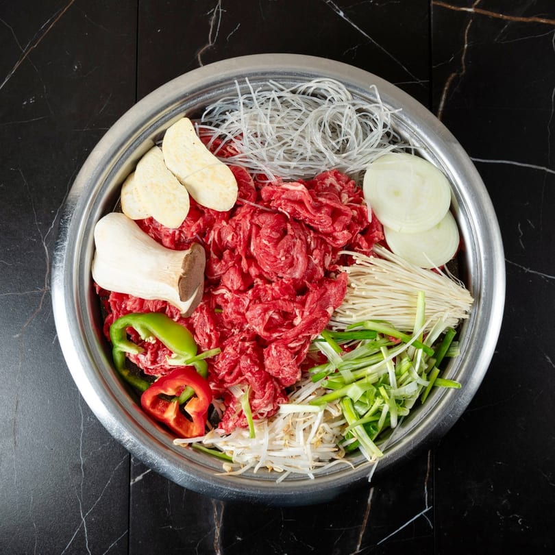 Overhead view of a metal bowl filled with raw beef, noodles, mushrooms, onions, and peppers.