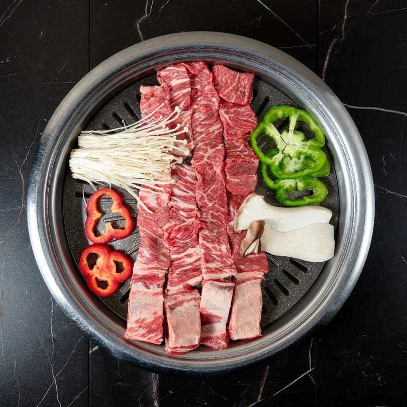 Raw beef short ribs, enoki mushrooms, bell peppers, and king oyster mushrooms on a grill.